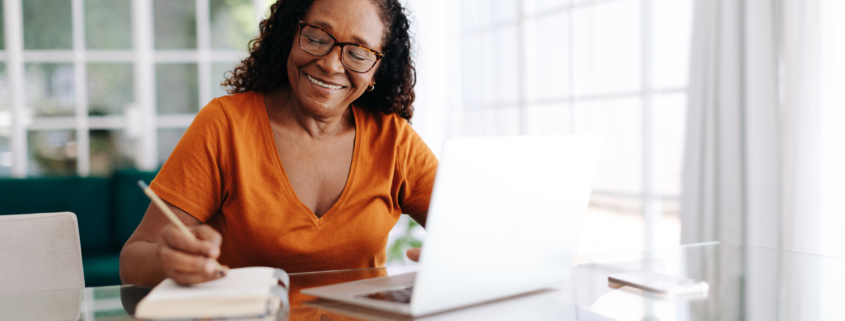 Happy senior woman sitting at a table in her home office, drafting her last will and testament in a journal and taking care to allocate her assets. Retired woman leaving a plan in place for the future.