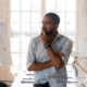 Pensive concerned african employee touch chin looking away leaned on table thinking inside of modern office boardroom, company owner boss search solution solve business issues or make decision concept